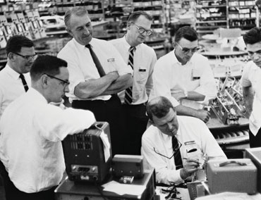 Dave Packard (standing with arms crossed) watches as Bill Hewlett takes a closer look at some HP instruments.